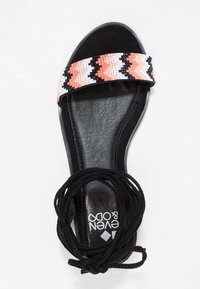 Flat sandal with a black leather sole, featuring a woven multi-color strap in pink, white, and black. Black lace wraps around the ankle.