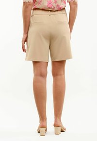 Γυναίκα με μπεζ tailored σορτς, φλοράλ κοντομάνικο πουκάμισο και μπεζ σανδάλια με χοντρό τακούνι, στέκεται με την πλάτη στην κάμερα.
