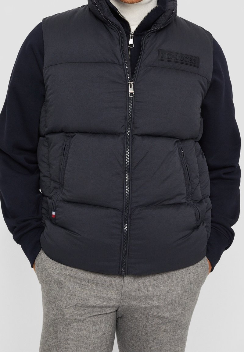 Navy Pufferveste mit hohem Kragen, Reißverschluss und Seitentaschen. Verfügt über ein erhabenes Logo-Patch auf der Brust und eine glatte, weiche Textur.