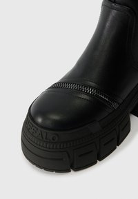 Bota de pele preta com sola de trato robusto, detalhe de fecho visível ao longo do dedo do pé, e a marca "BUFFALO" na borda da sola da frente.