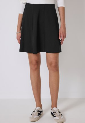 Jupe évasée noire en tissu doux, dotée de coutures. Assortie à des baskets blanches et beiges avec des accents et des lacets noirs.