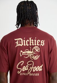 T-shirt en coton bordeaux avec du texte blanc et des graphiques d'un homard, avec les mots 'SeaFood' et 'Lunch-Dinner' imprimés dans une police ludique.