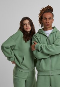 Deux personnes portent des sweats à capuche zippés en mint vert assortis, fabriqués en tissu doux, avec des poches avant et des poignets côtelés.