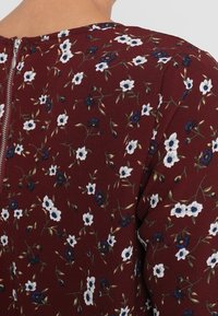 Tessuto bordeaux con un motivo floreale che presenta piccoli fiori bianchi e blu. Scollatura rotonda con una cerniera visibile sul retro.