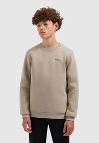 Tiener met krullend haar in een beige sweatshirt en zwarte broek, staande tegen een effen lichtgrijze achtergrond.