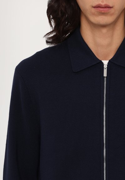 Casaco de malha azul-marinho com fecho zip, apresentando um colarinho, um fecho prata e textura em cordas. Design simples sem padrões ou acentos visíveis.