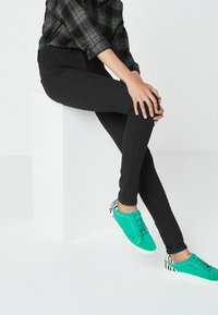Černé slim-fit kalhoty v kombinaci s tyrkysovými teniskami s akcenty zebřího vzoru. Oblečení má uvolněný, moderní vzhled s kostkovanou halenkou.