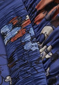 Stof met een bloemenpatroon op een diepblauwe basis, met geplooide textuur en gladde delen. Accenten van oranje en lichtblauw zichtbaar.