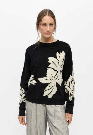 Femme portant un pull noir avec de grands motifs de feuilles blanches, associé à un pantalon ample gris clair, debout sur un fond blanc.
