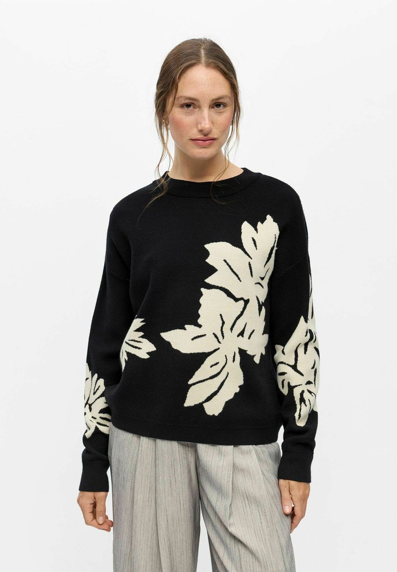 Femme portant un pull noir avec de grands motifs de feuilles blanches, associé à un pantalon ample gris clair, debout sur un fond blanc.