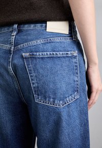 Blå denimjeans med rak passform, med en bakficka och en läderetikett märkta "CITIZENS of HUMANITY" nära midjebandet.