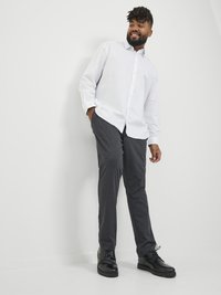 Camisa blanca de botones, pantalones gris oscuro y zapatos negros de cordones. Corte ajustado con textura suave. De pie frente a un fondo sencillo.