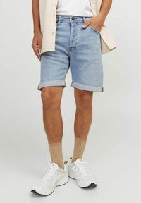 Man wearing light blue rolled-up denim shorts, beige ribbed socks, white sneakers, and a beige shirt with one hand in pocket.