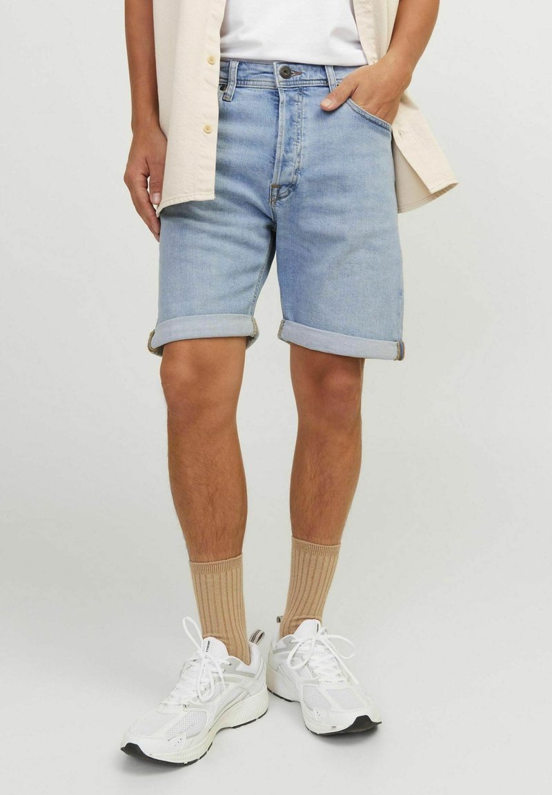 Man wearing light blue rolled-up denim shorts, beige ribbed socks, white sneakers, and a beige shirt with one hand in pocket.