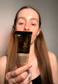 Jong persoon met lang haar houdt een tube hydraterende make-up dicht bij het gezicht tegen een effen achtergrond.