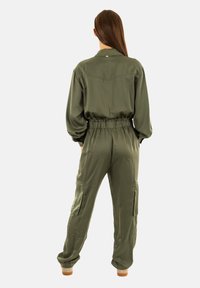 Jumpsuit verde oliva realizzato in tessuto morbido, con maniche lunghe, vita elasticizzata, tasche cargo e colletto.