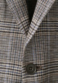 Bruine, beige en blauwe geruite colbertkraag met een grote donkerbruine knoop die aan de voorkant is vastgemaakt.