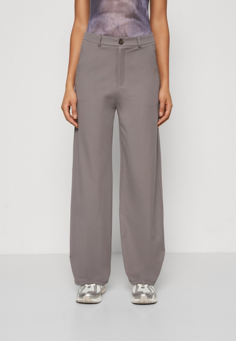 Hoge taille, wijde broek van zacht, grijs stof; heeft een sluiting met een knoop aan de voorkant en een strakke, minimalistische uitstraling. Draagt met grijze sneakers.