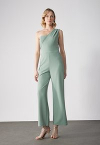 Mono de una sola manga en tela verde claro; cuerpo ajustado y diseño de pierna ancha; textura suave; tacones dorados con tiras como calzado.