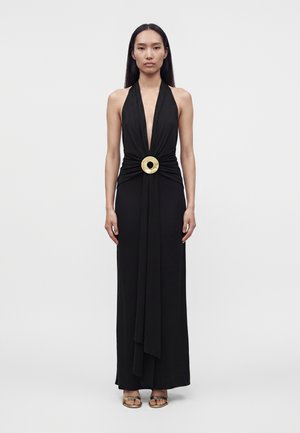 Frau trägt ein ärmelloses schwarzes Kleid mit Halterneck und tiefem V-Ausschnitt sowie einer großen runden goldenen Schnalle an der Taille, steht vor einem einfarbigen Hintergrund.