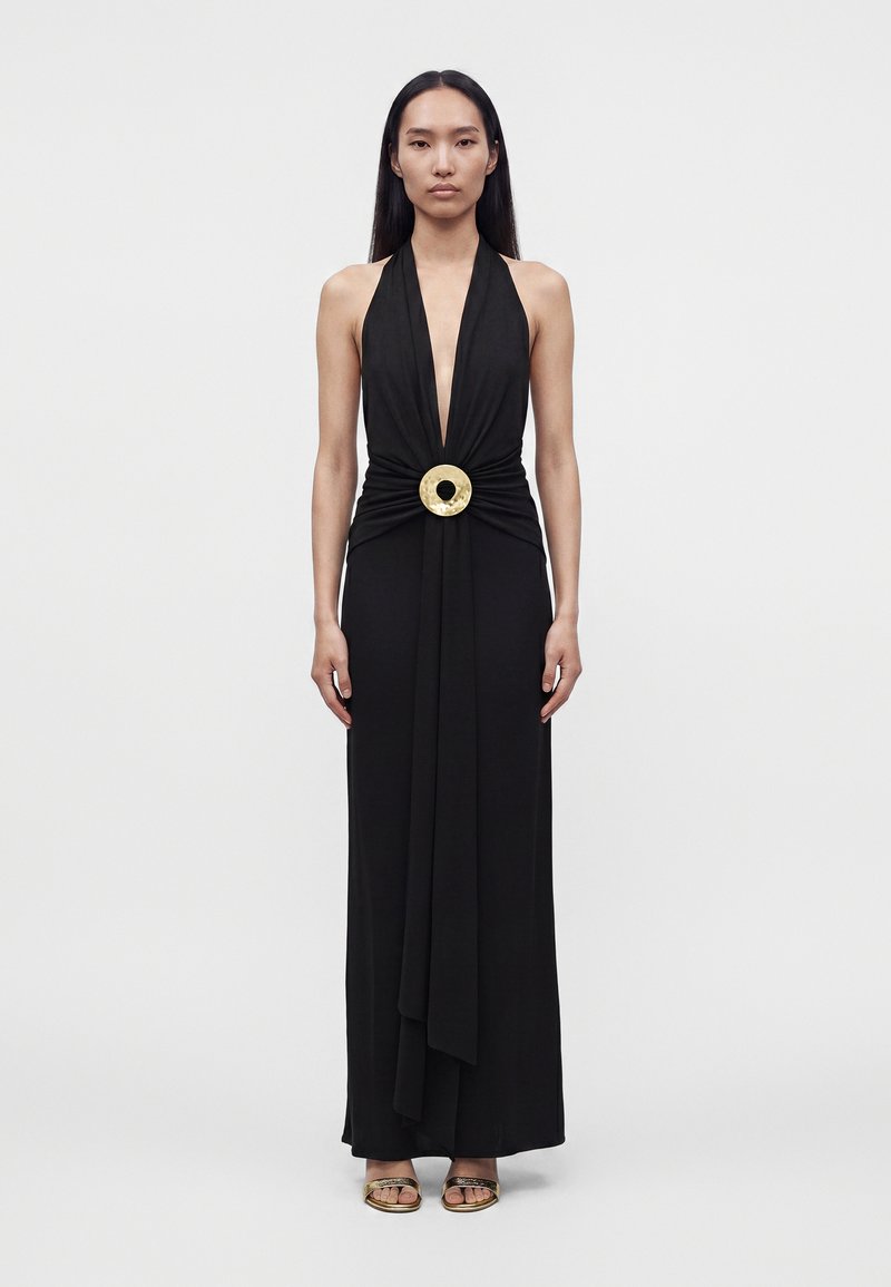 Woman wearing a sleeveless black halter-neck gown with a deep V and a large circular gold buckle at the waist, standing against a plain background.