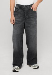 Jeans a gamba larga in denim nero con una finitura sbiadita, tasca anteriore singola e dettagli di cuciture a contrasto. Indossati con sneakers grigie.