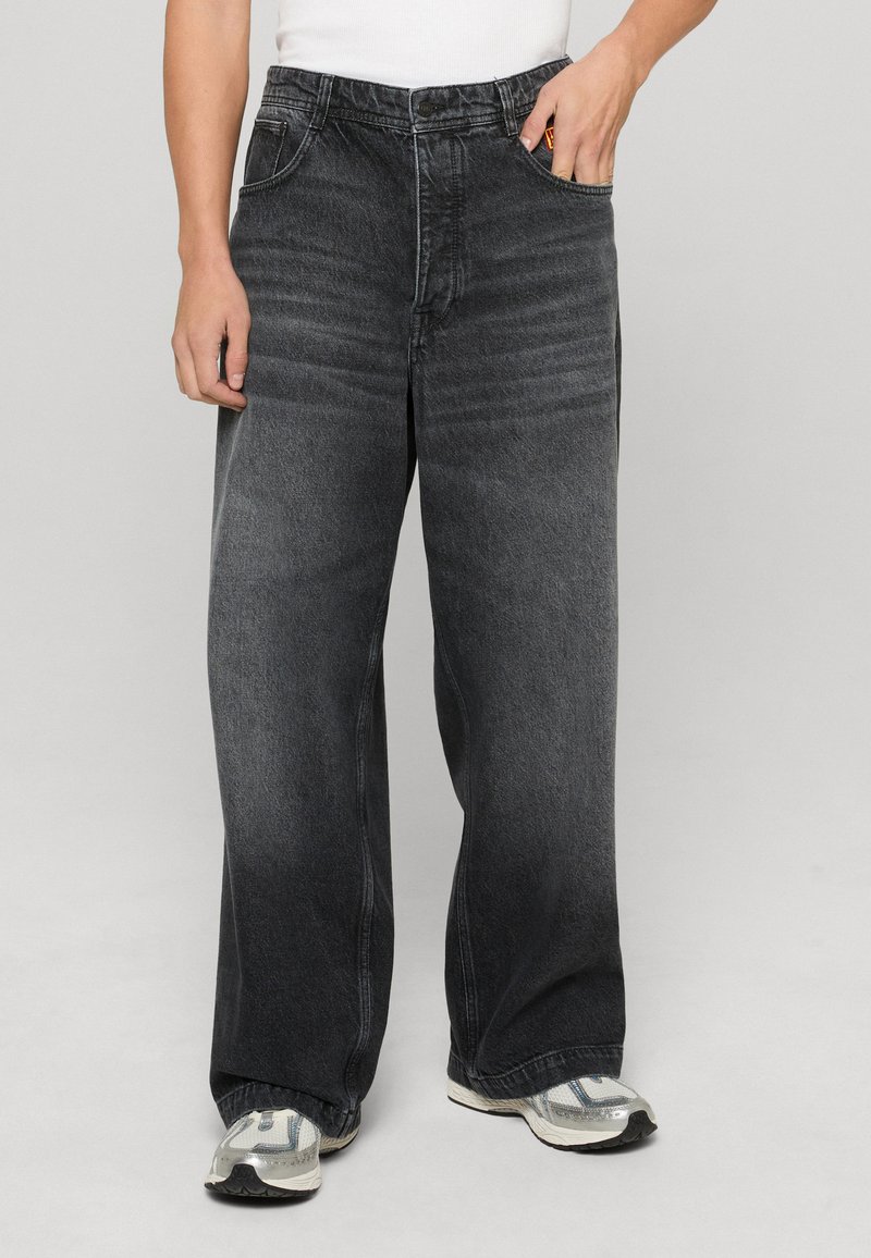 Jeans a gamba larga in denim nero con una finitura sbiadita, tasca anteriore singola e dettagli di cuciture a contrasto. Indossati con sneakers grigie.