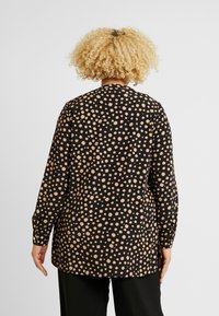 Maglietta nera a maniche lunghe con motivo a pois beige, con taglio dritto e polsini con bottoni. Tessuto morbido, vestibilità ampia e spacchetti laterali.