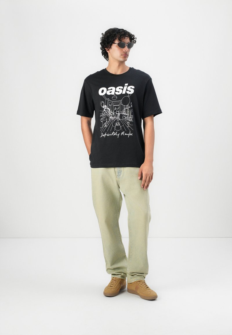 Sort grafisk t-shirt med "oasis" og linjeillustration; parret med lysegrønne bukser og brune sneakers. Runde solbriller fuldender looket.