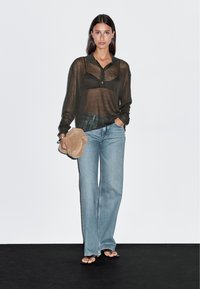 Schwarze durchsichtige Langarmbluse mit Kragen, hellblaue Schlagjeans, hält ein beigefarbiges Täschchen. Kombiniert mit schwarzen Flip-Flops. Weißer Hintergrund.