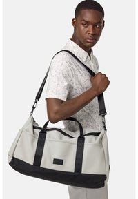 Borsa duffel sintetica grigia chiara con accenti neri, dotata di manici doppi e di una tracolla regolabile, progettata per versatilità e durevolezza.