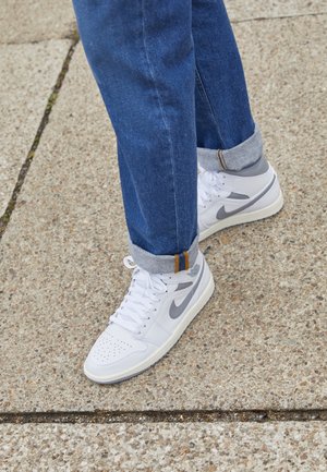 Person trägt hochgekrempelte blaue Jeans und weiße Nike-Sneakers und steht auf einem strukturierten Betonboden.
