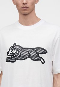 Mann trägt weißes T-Shirt mit großem schwarzem Motiv eines laufenden Tieres, das aus kleinen Pfotenabdrücken besteht, auf der Vorderseite.