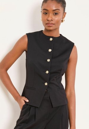 Frau trägt ärmellose schwarze, maßgeschneiderte Weste mit goldenen Knöpfen und passende schwarze Hose, posiert mit einer Hand in der Tasche.