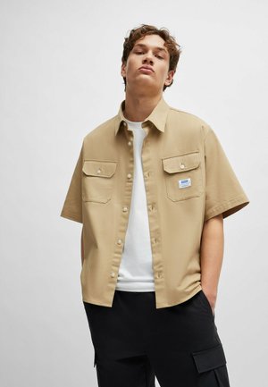 Short-sleeve, beige button-up shirt with two chest pockets and a branded patch. Worn over a white T-shirt. Smooth fabric texture.