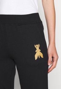 Svarta joggers i mjukt tyg med en gyllene broderad design av en figur på vänster sida. Elastisk midjebands synlig.