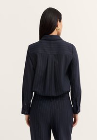 Navy pinstriped blouse with a collar, shoulder pleats, and buttoned cuffs. Made of smooth fabric, featuring subtle golden thread accents.