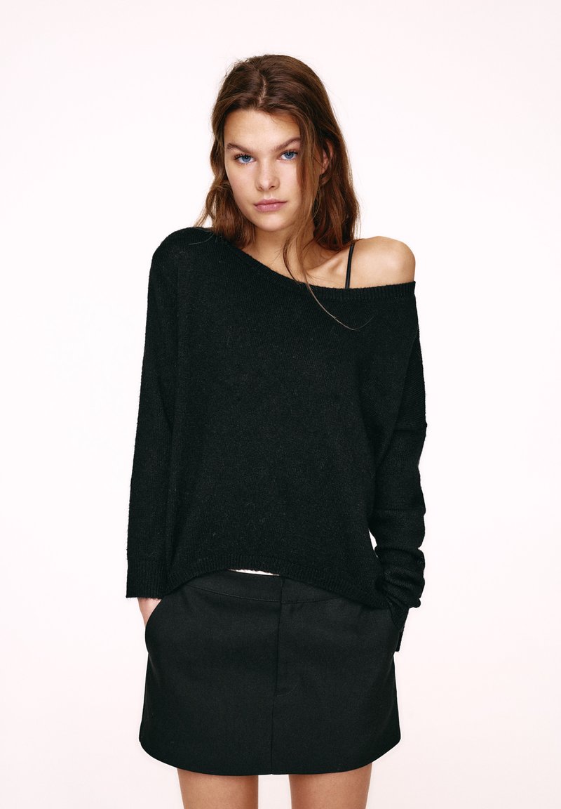Pull noir à mailles larges avec un design épaules dénudées, manches larges et texture douce, associé à une jupe noire.