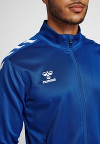 Blue zip-up jacket made of smooth fabric, featuring a high collar and white Hummel logo with chevron accents on the shoulders.