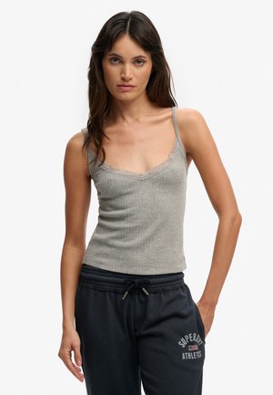 Femme portant un caraco gris orné de dentelle et un pantalon de survêtement noir Superdry Athletics, debout avec une main dans la poche.