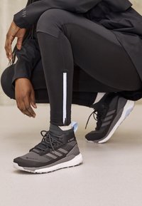 Persona vestida con leggings deportivos negros con cremallera blanca y zapatillas Adidas Terrex Gore-Tex grises, agachada sobre un suelo claro.