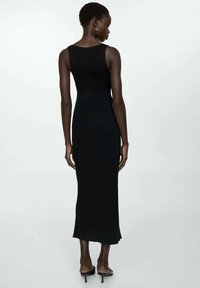 Robe maxi en tricot côtière noire avec une silhouette ajustée, décolleté dégagé et une fente subtile à l'ourlet. Portée avec des chaussures à talons fins.