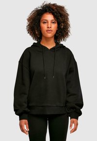 Sudadera negra con capucha, de corte holgado, puños y dobladillo acanalados, capucha con cordón, y textura de felpa suave. Se lleva con leggings negros.