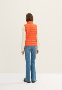 Gepolstertes oranges Weste mit hohem Kragen, getragen über einem leichten Pullover. Die Rückansicht zeigt vertikale Nähte und eine enge Passform, kombiniert mit blauen Schlagjeans.