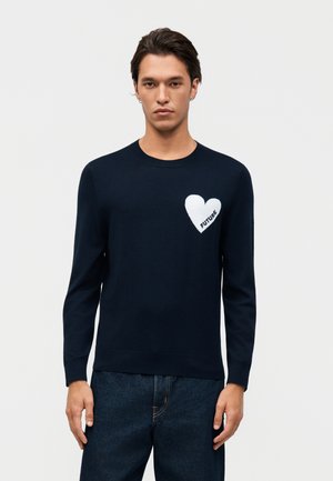 Jeune homme portant un pull à manches longues noir avec un cœur blanc et le mot « FUTURE » sur le côté gauche de la poitrine, associé à un jean foncé.