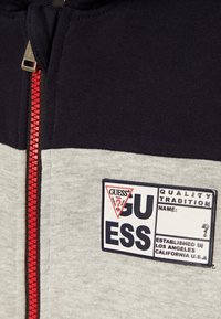 Hoddie zippé gris et marine avec une fermeture éclair rouge, comportant un patch logo rectangulaire avec des détails de marque. Texture de tissu lisse et design décontracté.