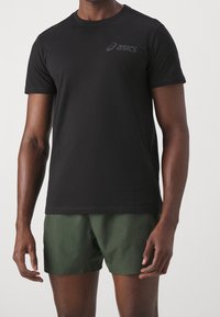 Camiseta negra de manga corta hecha de algodón, con un sutil logo gris en el pecho. Combinada con pantalones cortos verdes que tienen una textura suave.