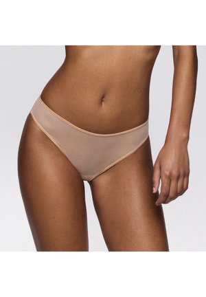 Femme portant un sous-vêtement bikini sans coutures couleur chair, vue de la taille à mi-cuisse sur un fond clair uni.
