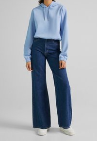 Sudadera con capucha azul claro con cordones, combinada con vaqueros azul oscuro de cintura alta y pierna ancha, y zapatillas blancas. Texturas suaves y diseño casual.