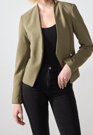 Frau trägt olivgrünen Reißverschlussblazer über schwarzem Top und schwarzen hoch taillierten Jeans, Hand ruht nahe der Hüfte mit Goldringen.